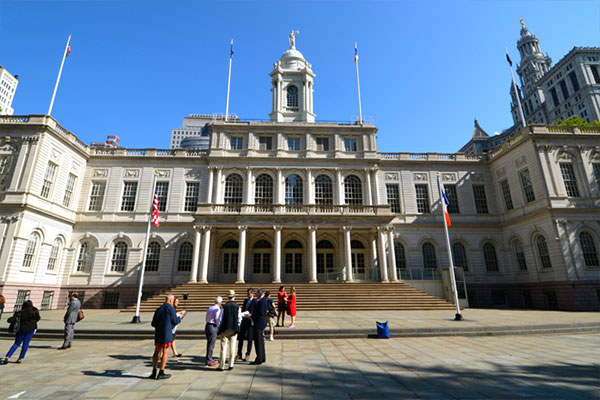 Front view of a grand neoclassical city hall-style building with a central clock tower, flags, and steps with people on the plaza