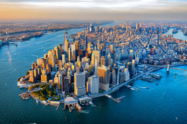 Aerial view of Manhattan's dense skyline with the East River and bridges at sunset, showing tall skyscrapers along the waterfront.